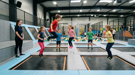 Des enfants sautent sur les trampolines d'un parc indoor pendant qu'un encadrant surveille depuis la plateforme.